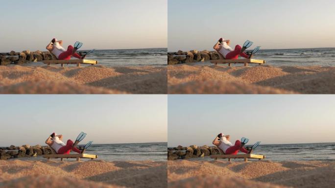圣诞老人日光浴。戴着墨镜和脚蹼的有趣的圣诞老人坐在躺椅上，在海边的海滩上，手持小型音乐扬声器听音乐，
