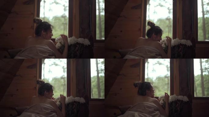 秋天的早晨，年轻迷人的亚洲女人躺在窗边的床上，看着木制小屋外美丽的大自然。微笑的漂亮女孩放松，享受假