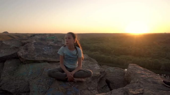 一个甜美沉思的少女坐在悬崖顶上，顶着黄色的夕阳。她的头发随风飘扬。日落时美丽的风景。肖像。4K。