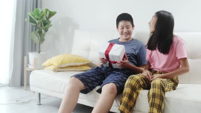 快乐的亚洲女孩给她的哥哥礼品盒，而他坐在家里的沙发上庆祝节日的概念。
