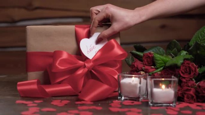 桌子上有两支蜡烛，一个包装好的盒子和一束玫瑰。女人的手拿一张带有爱这个词的心形情人节卡片