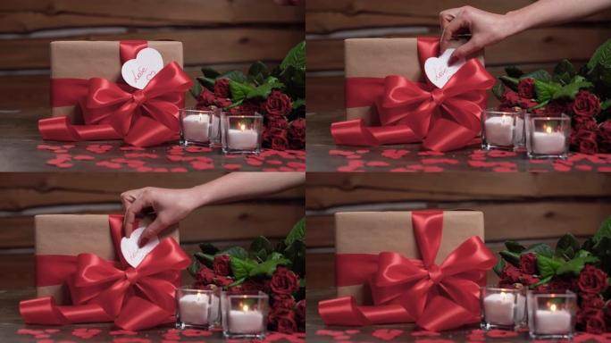 桌子上有两支蜡烛，一个包装好的盒子和一束玫瑰。女人的手拿一张带有爱这个词的心形情人节卡片