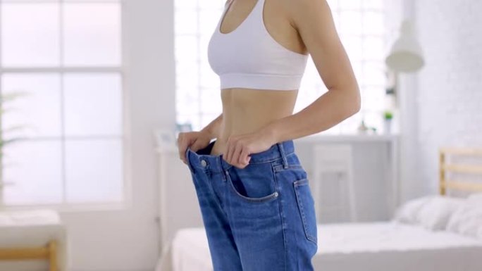 穿着大牛仔裤的年轻苗条女人在家里展示她的饮食效果