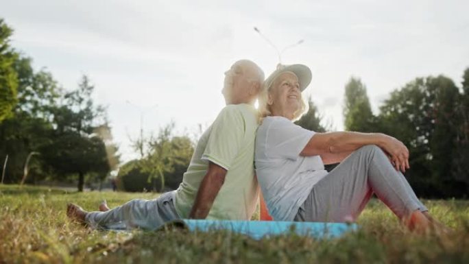 穿着运动服的老年男女在公园健身锻炼后背靠背坐在草地上微笑的侧视图