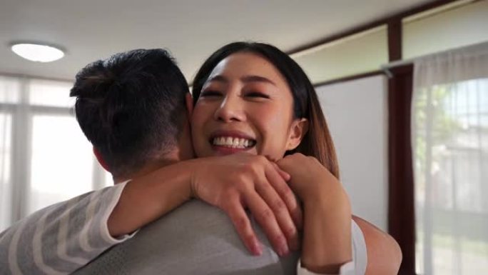 30多岁的亚洲年轻人在家里的客厅里跑去拥抱一个微笑的女人。幸福的夫妻拥抱并在舒适的房子里度过家庭时光