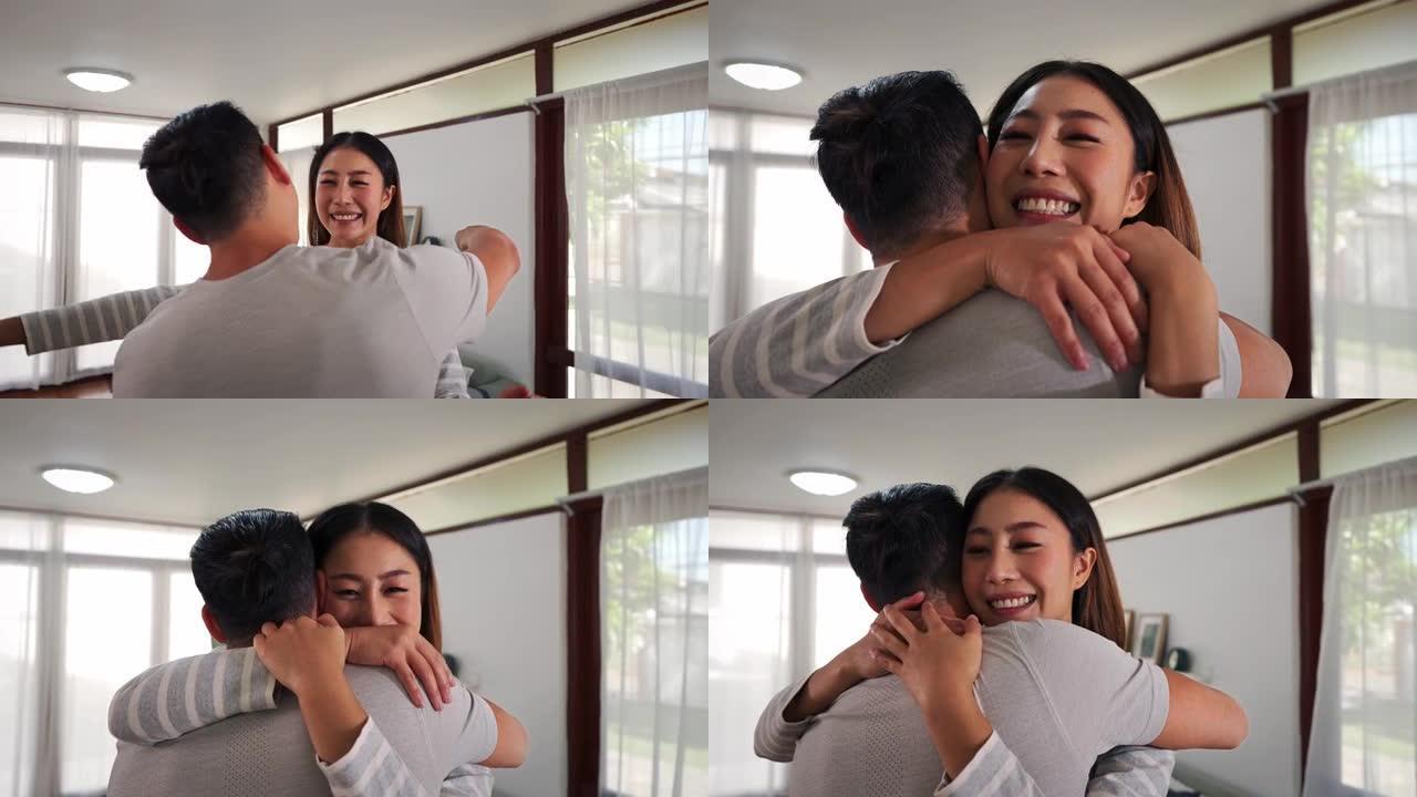 30多岁的亚洲年轻人在家里的客厅里跑去拥抱一个微笑的女人。幸福的夫妻拥抱并在舒适的房子里度过家庭时光