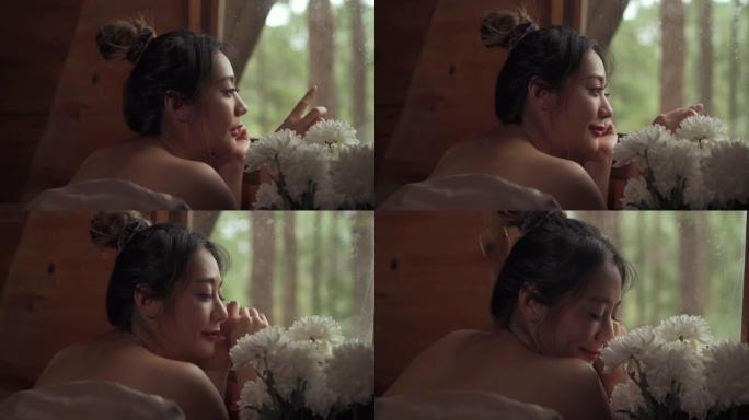 秋天的早晨，年轻迷人的亚洲女人躺在窗边的床上，看着木制小屋外美丽的大自然。微笑的漂亮女孩放松，享受假