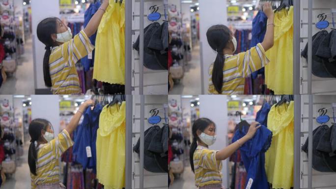 戴着面具的亚洲女孩在购物中心选择衣服，新型冠状病毒肺炎期间的新常态概念。