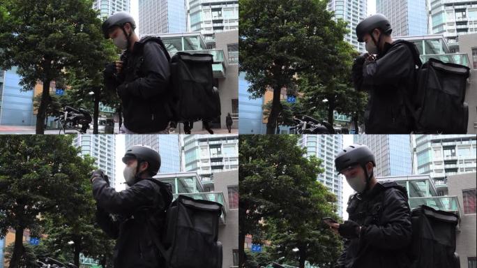 戴着黑色背包和防护面罩的年轻人快递员手里拿着电话，环顾四周。自行车快递员寻找地址。在手机上使用导航应