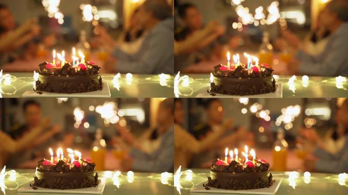 为儿童生日聚会准备的桌子上配有烛光的巧克力蛋糕。家庭中的人在后台玩烟火。