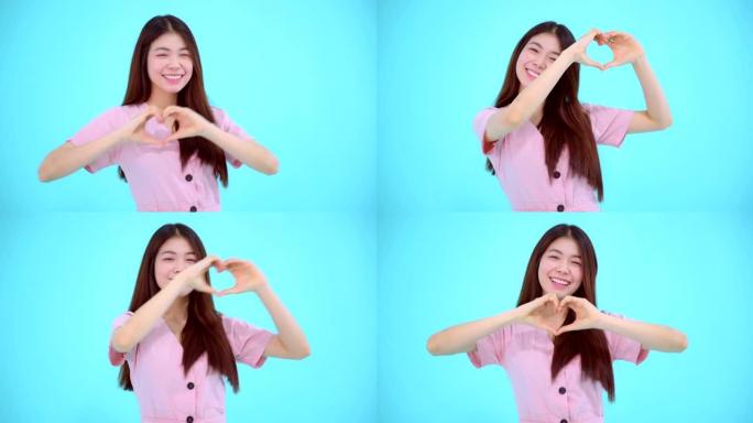 亚洲女人在爱情中快乐微笑，用手塑造心形，看着孤立在蓝色背景上的相机。4k视频。快乐的情感概念。
