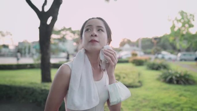 亚洲活跃的年轻女子在公园散步，模特擦着出汗的脸白毛巾，边锻炼边听音乐，人寿保险计划，健康的生活方式，