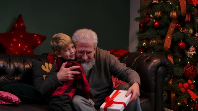 可爱的小男孩给他的祖父一个很棒的圣诞夜礼物。一位年迈的祖父在一棵发光的圣诞树附近的装饰房间里拥抱他的