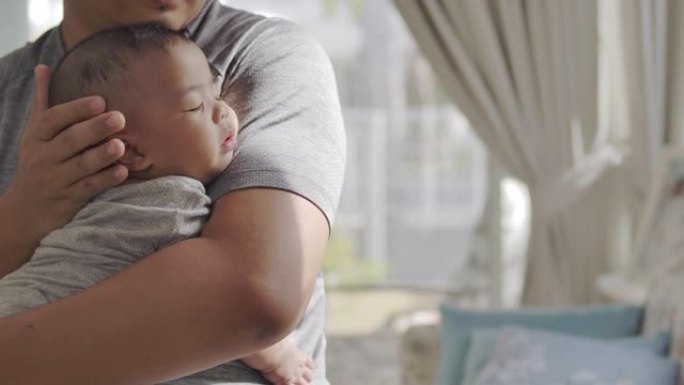 一位年轻的亚洲全职父亲抱着并安抚婴儿，在爱与关怀的陪伴下在客厅睡觉。