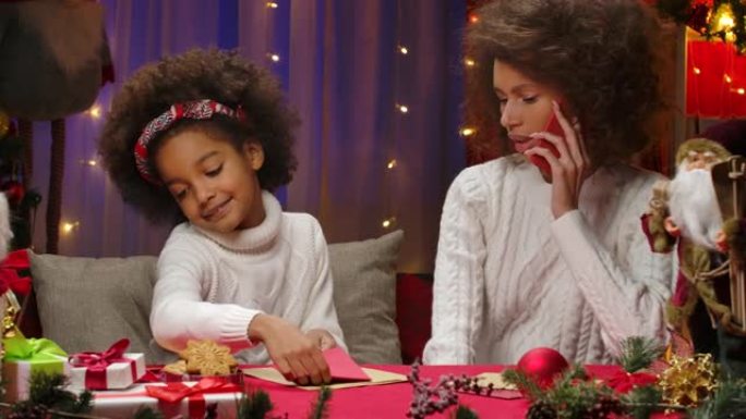 非裔美国妇女在智能手机上聊天。妈妈和女儿穿着白色毛衣坐在家庭房间的背景下，为圣诞节假期装饰。特写