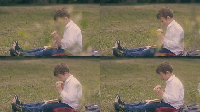金发的饥饿男孩坐在草坪上吃大汉堡