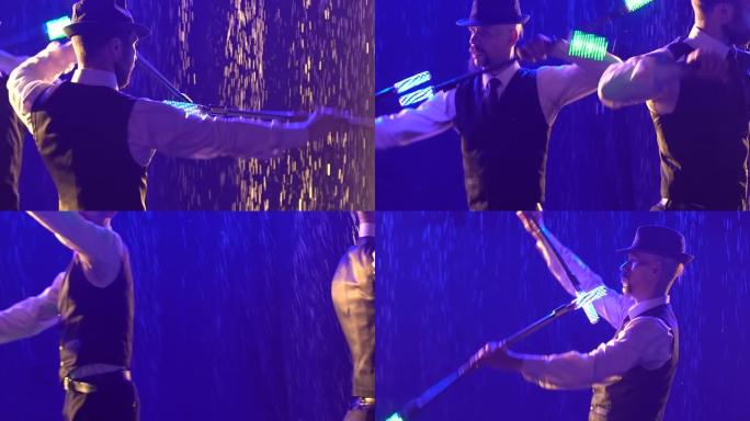 两个戴着帽子的时髦男人在雨滴下慢动作表演的肖像。艺术家的特写剪影出现在旋转荧光棒的灯光下