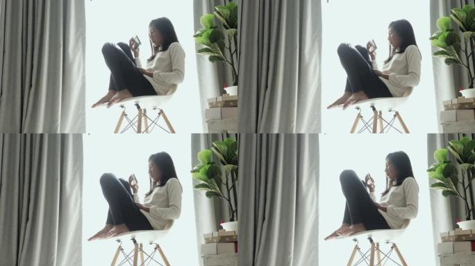亚洲女孩坐在椅子上，窗灯和使用智能手机在早上在家里，生活方式的概念。
