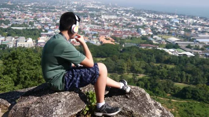 快乐的亚洲男孩在阳光明媚的日子里戴着耳机在山顶上听音乐，背景自然。