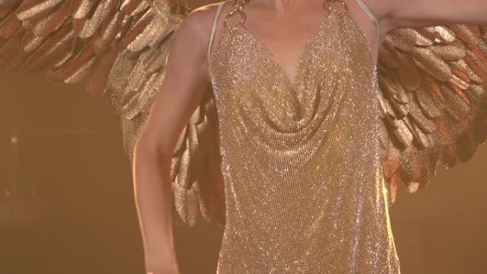 华丽的女星扮成带翅膀的希腊女神阿耳忒弥斯。一个穿着金色西装和头饰的苗条女人在工作室灯光的背景下跳舞。