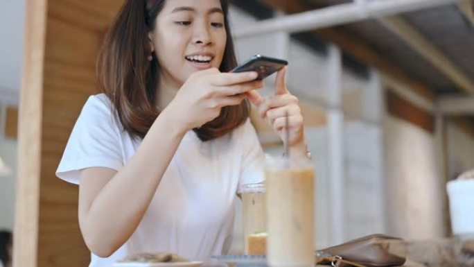 美丽的年轻亚洲女人喜欢在咖啡馆拍照，等她的朋友，喝咖啡，上传照片到社交网络微笑和开朗的脸真实的人