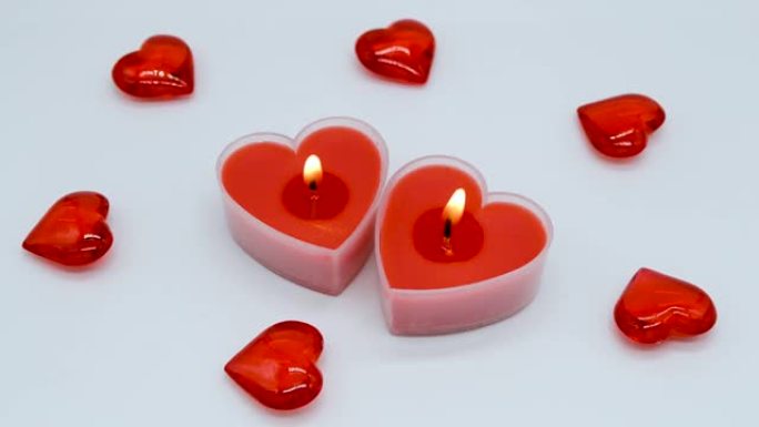模糊的框架变得清晰: 两支鲜红色的心形蜡烛在白色背景上燃烧，并装饰着小的玻璃心。蜡烛燃烧了一会儿，最
