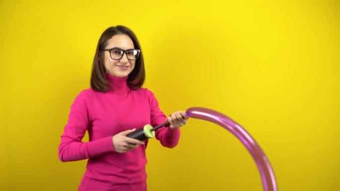 一名年轻女子用泵给一个长长的紫色气球充气，它在黄色背景上爆裂。穿着粉色高领毛衣和眼镜的女孩。