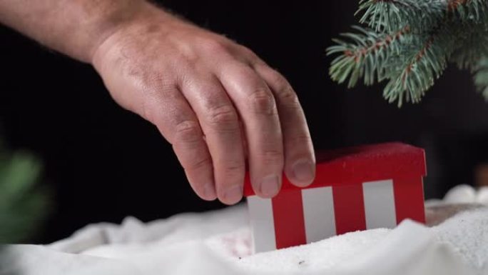 一个红色的盒子躺在云杉树枝下的白色雪地上。男性手打开它。圣诞礼物概念