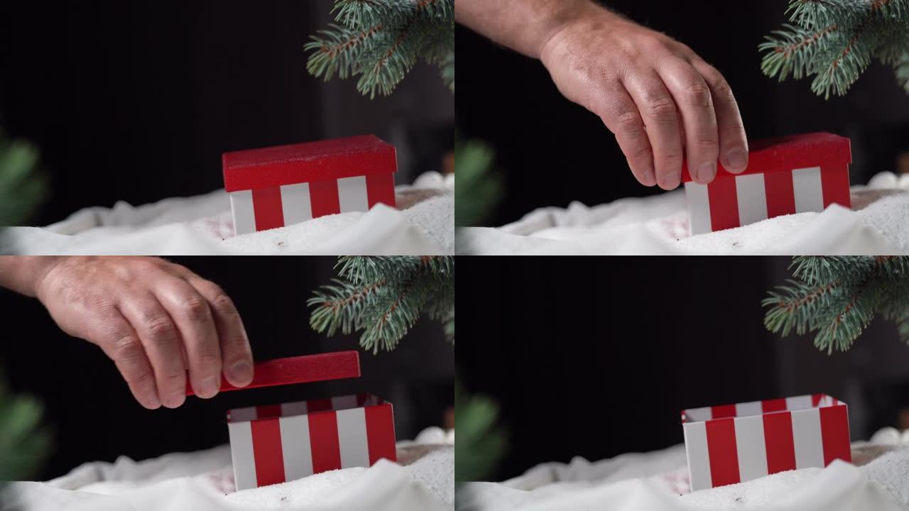 一个红色的盒子躺在云杉树枝下的白色雪地上。男性手打开它。圣诞礼物概念