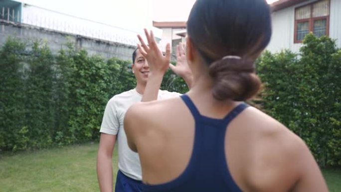 年轻的健身夫妇在房子的花园外面做高五手势。亚洲妇女和高加索男子在公园户外训练