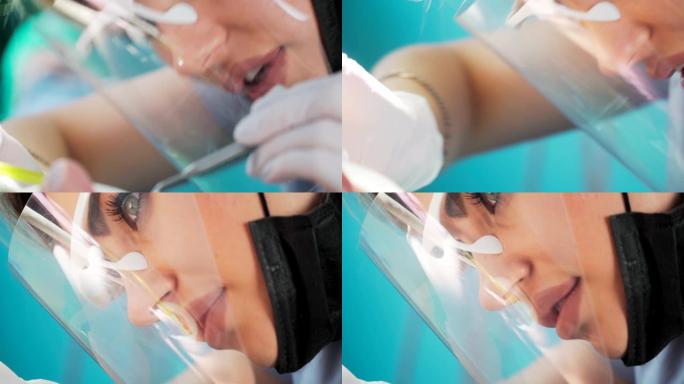 美丽的女人牙医在一个保护盾和口罩的帮助医疗设备与一名女性患者在牙科办公室工作。特写。
