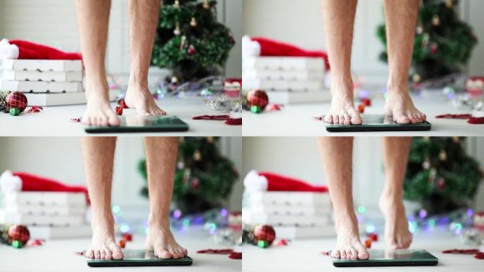 男性腿的特写站立在秤上测量体重。假期圣诞节过后，披萨盒和新年是装饰品，瓶子躺在地板上。人体测量身体脂