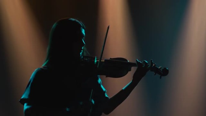 一个拉小提琴的女人的深色剪影。一位女音乐家在一个黑暗的工作室的背景下演奏，该工作室充满烟雾和舞台灯光