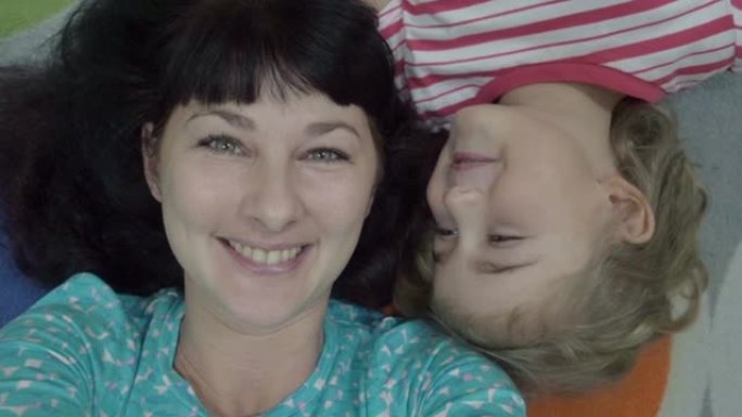 家庭自拍照。兴奋的年轻母亲和孩子躺在地板上玩耍，自拍。母女俩在镜头前四处闲逛并接吻。妈妈和宝宝特写的