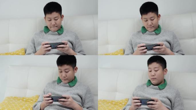 亚洲青春期男孩穿着灰色毛衣坐在沙发上，在家通过智能手机玩网络游戏，生活方式概念。