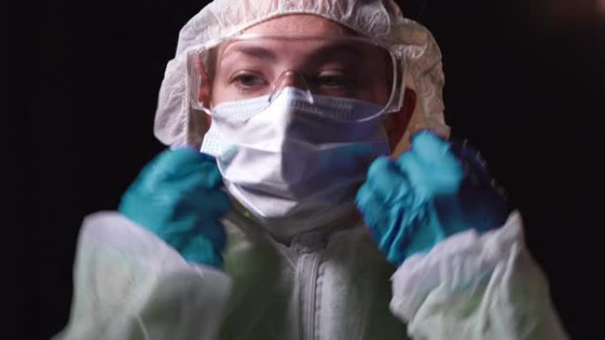 年轻的女医生穿着白色防护服，戴着眼镜，戴着医用口罩站在黑色背景上，整理衣服。医生回收概念