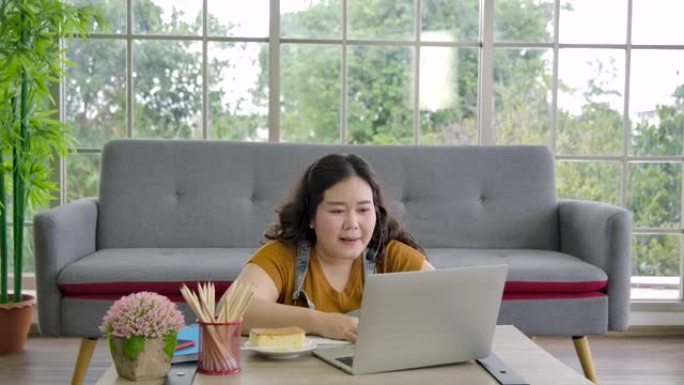 超重的女商人在家使用笔记本电脑。同时乐于吃零食。