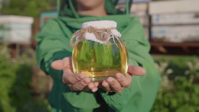 带蜂蜜的养蜂人。一位女农民从养蜂场展示蜂蜜。玻璃罐子里的黄色蜂蜜