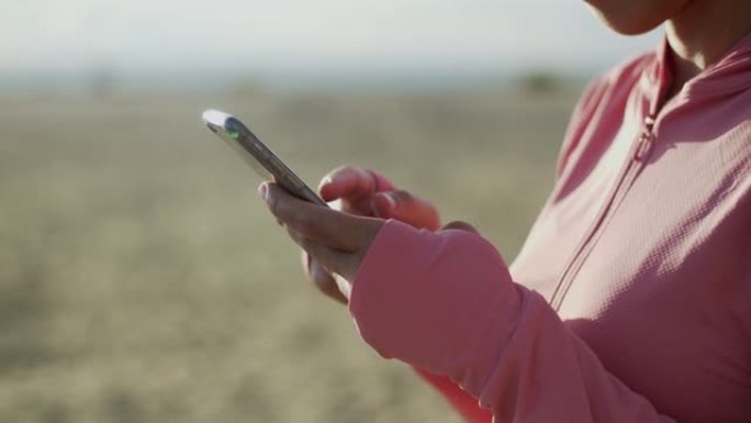 在早晨日出时站在海滩上的一名女孩体育运动员在智能手机上近距离输入一条消息。健康的生活方式观念。