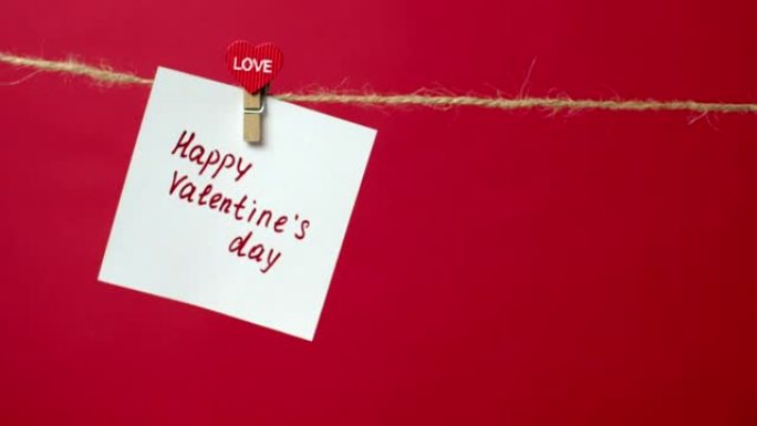 绳子上的纸条特写，上面写着情人节快乐。女孩的手将一张白纸挂在衣夹上的红色背景上。爱情宣言、浪漫关系的