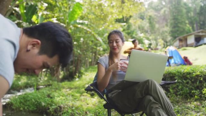 美丽的亚洲女人在森林营地使用笔记本电脑和互联网在户外工作的慢动作。幸福的夫妇坐在小溪边的椅子上，一起