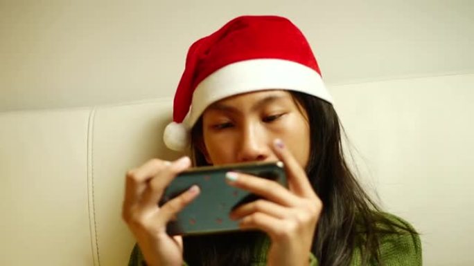 快乐的亚洲女孩穿着红色圣诞老人帽子绿色毛衣，在圣诞节假期在家沙发上玩网络游戏。