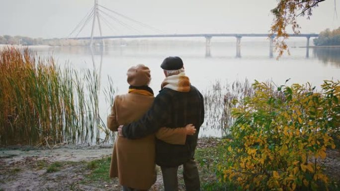 充满爱心的老年夫妇拥抱和微笑，站在河岸上，看着巨大的桥梁穿越它，秋天