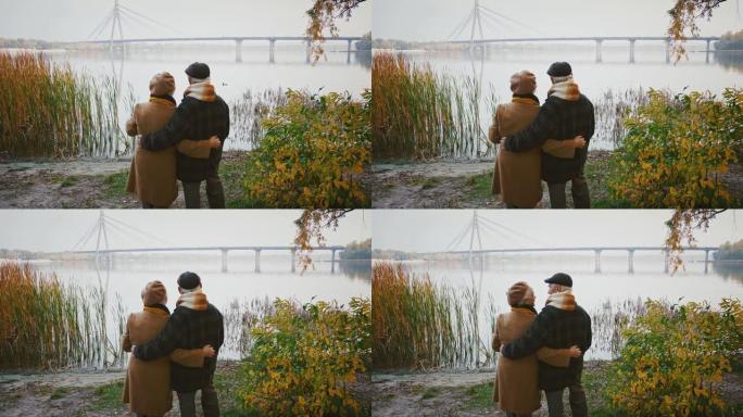 充满爱心的老年夫妇拥抱和微笑，站在河岸上，看着巨大的桥梁穿越它，秋天