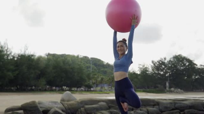 快乐的亚洲女人在海边的瑜伽垫上展示健身球