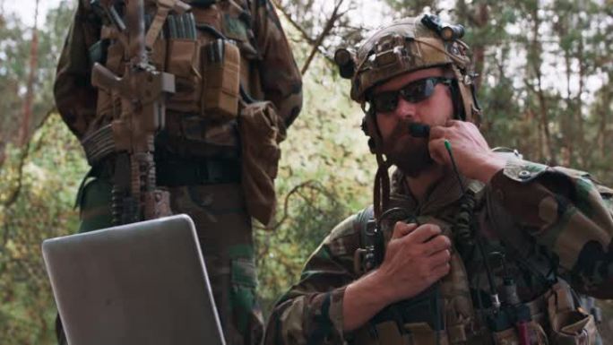 这是一幅身着军装、身穿战术背心、头戴头盔的指挥官肖像，他在笔记本电脑的键盘上打字，通过军用无线电报告
