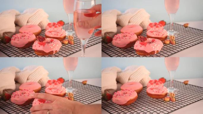 烤架上有两杯粉红色的葡萄酒或香槟和粉红色的甜甜圈。情人节概念。女人手拿一个杯子和甜甜圈