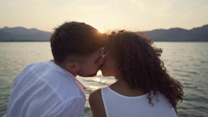 一对多种族的夫妇在背光中亲吻，湖中夕阳西下，脸上有光线反射-两个美丽的异性恋恋人交换了来自背后的感情