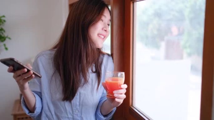 可爱的少女或女人的肖像用手机在玻璃窗前喝橙汁，在家看起来很开心