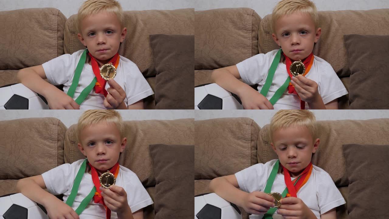 一个小男孩坐在沙发上，拿着足球和很多奖牌。